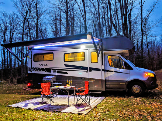 alt= Recreational vehicle parked in a campsite at dusk with awning extended and a cozy outdoor seating arrangement.